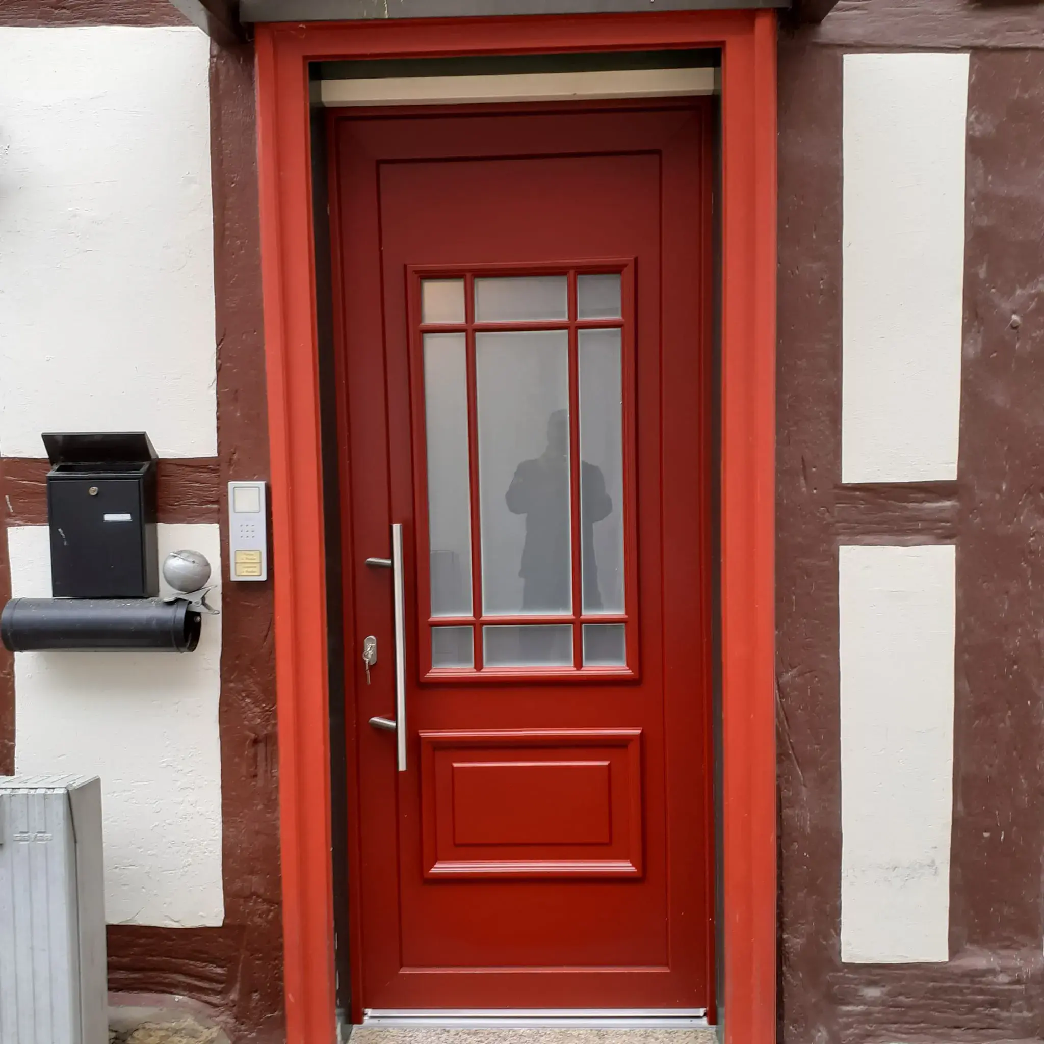 Rote Haustür mit Glasfenster, links Briefkasten und Klingel, Fachwerkhauswand.