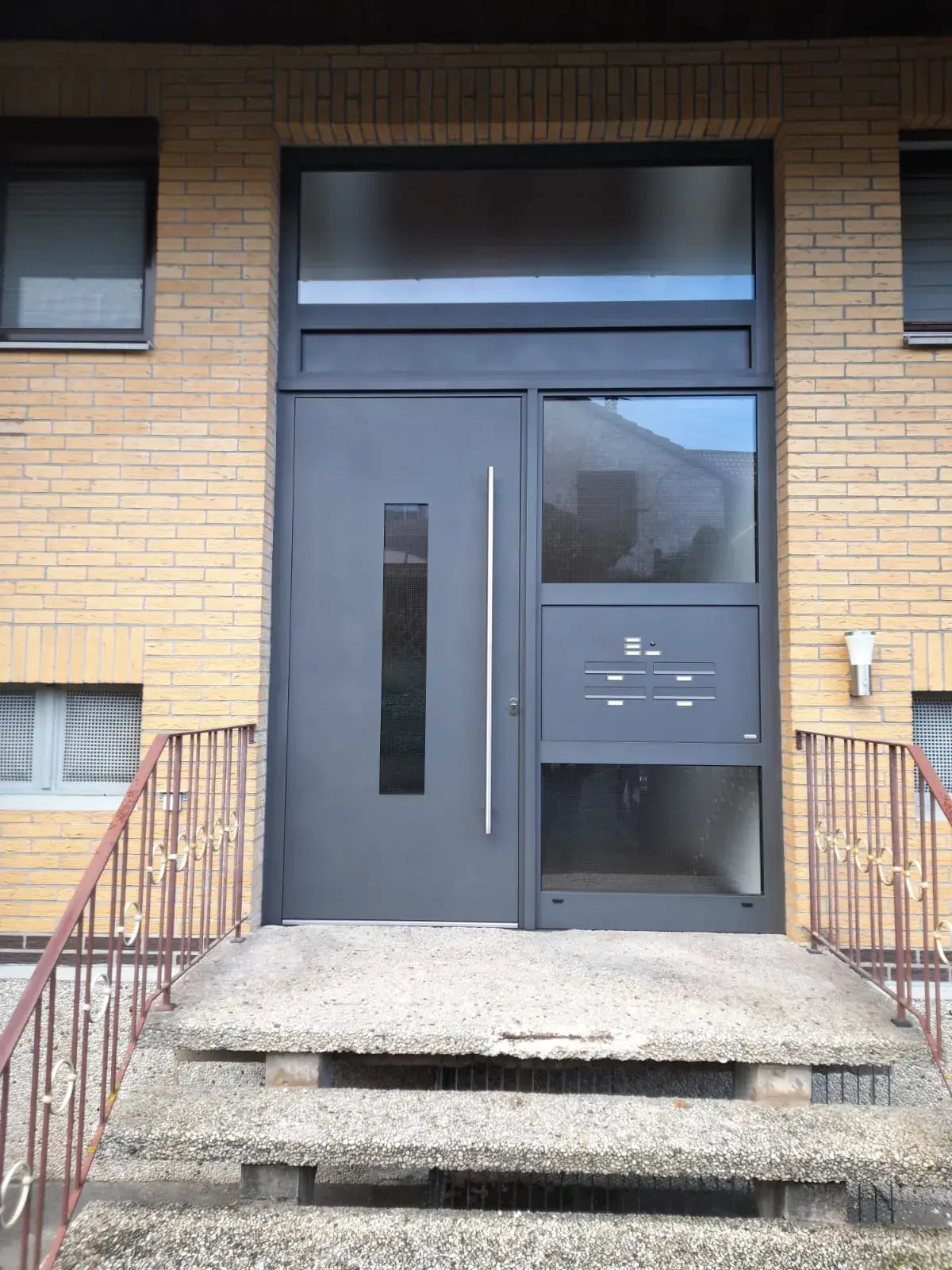 Moderne graue Haustür mit Briefkästen, Treppe und gelber Klinkerfassade.