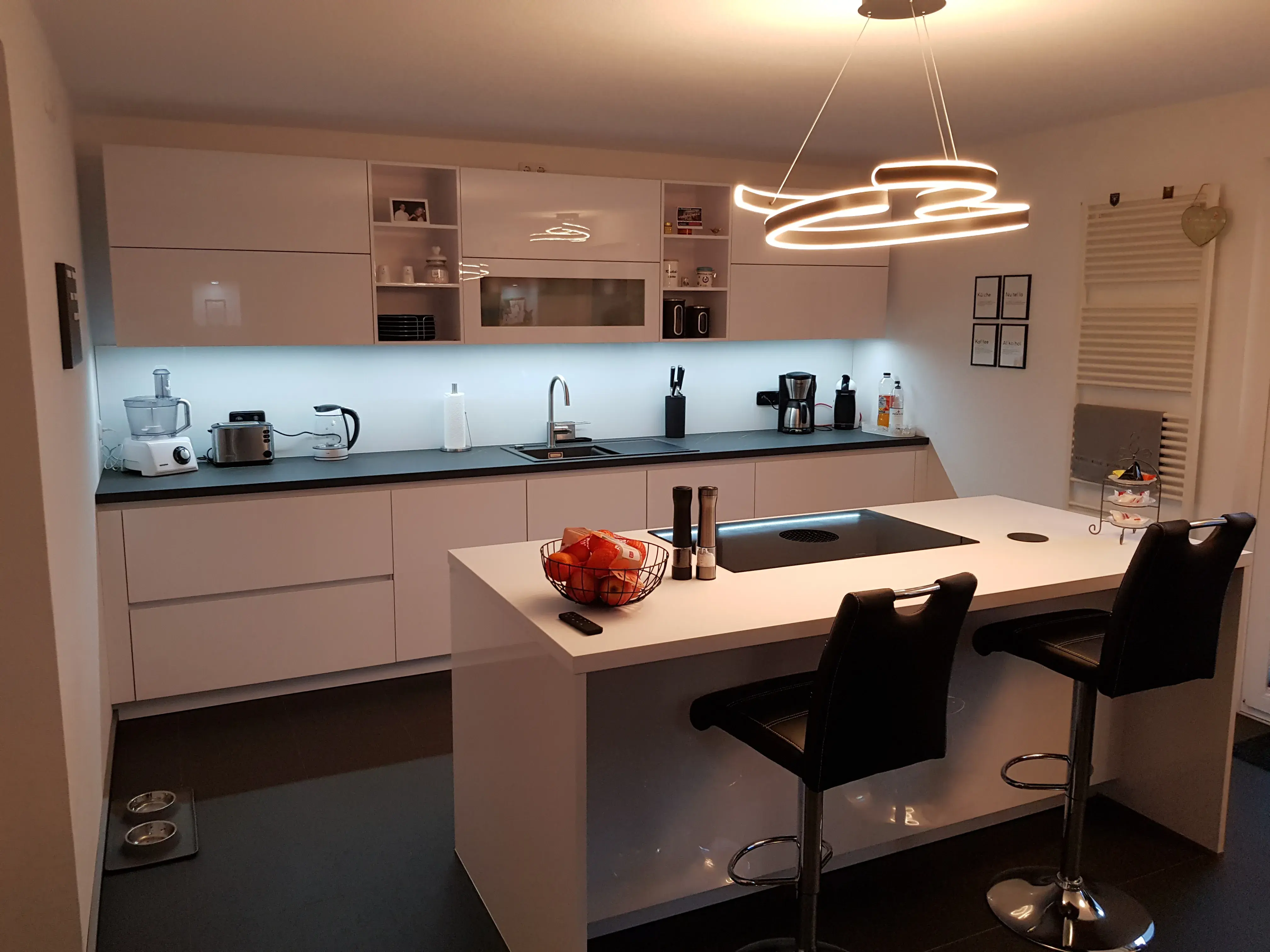 Moderne weiße Küche mit Insel, zwei Barhockern und heller LED-Beleuchtung.
