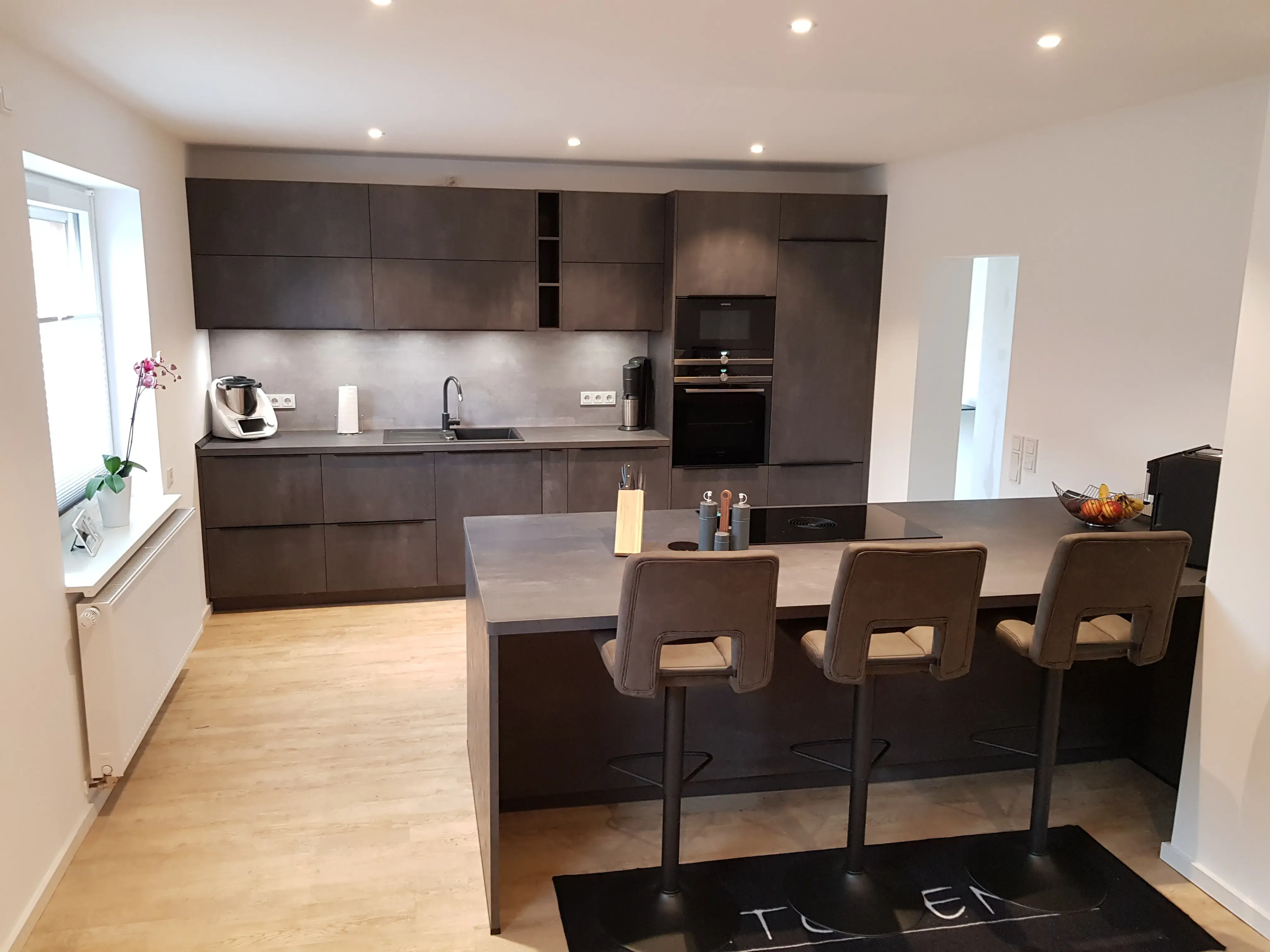 Moderne Küche mit dunklen Schränken, Kochinsel und drei Barhockern, hell beleuchtet.