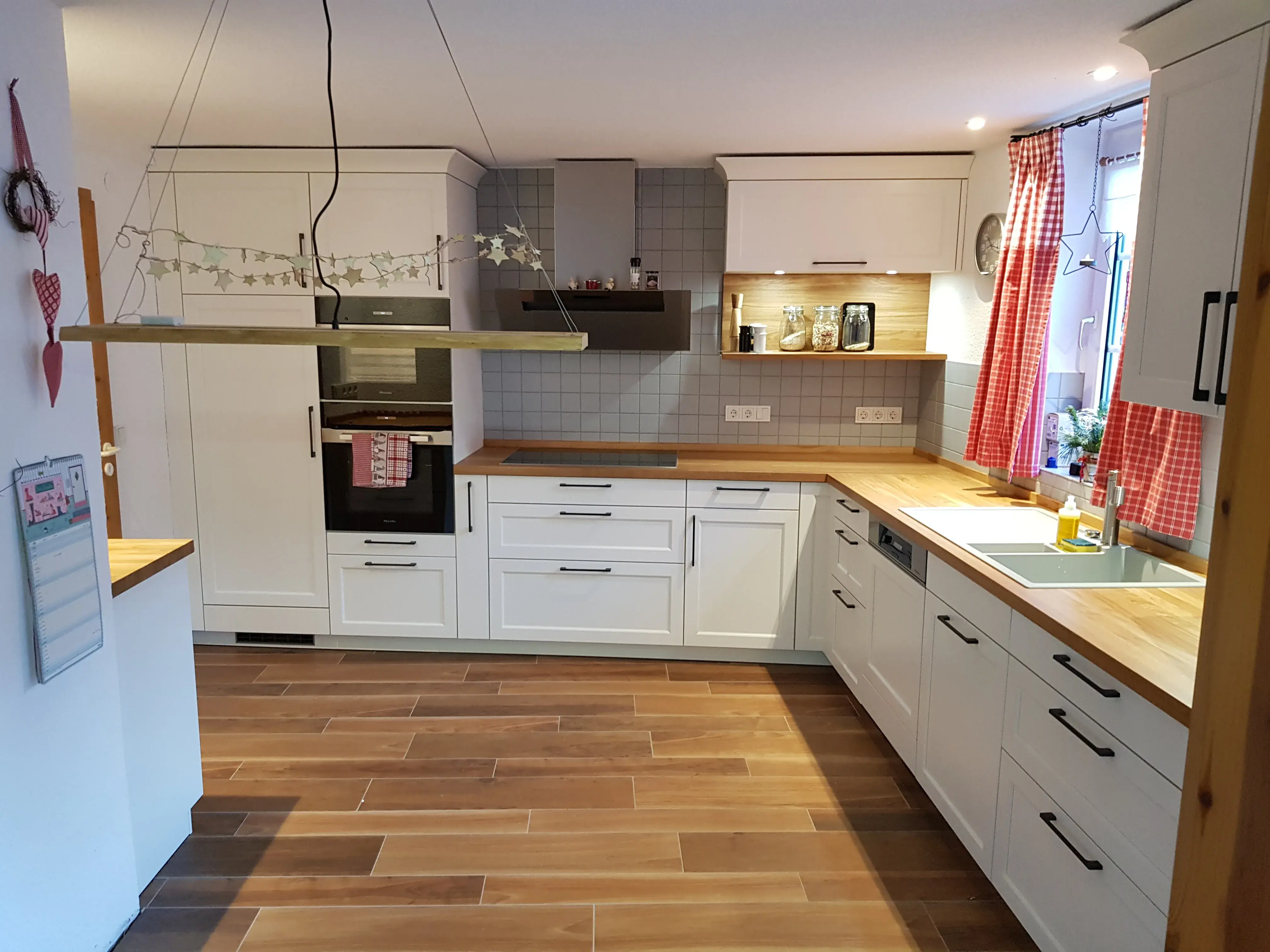 Helle Küche mit weißen Schränken, Holzarbeitsplatten und roten Vorhängen am Fenster.