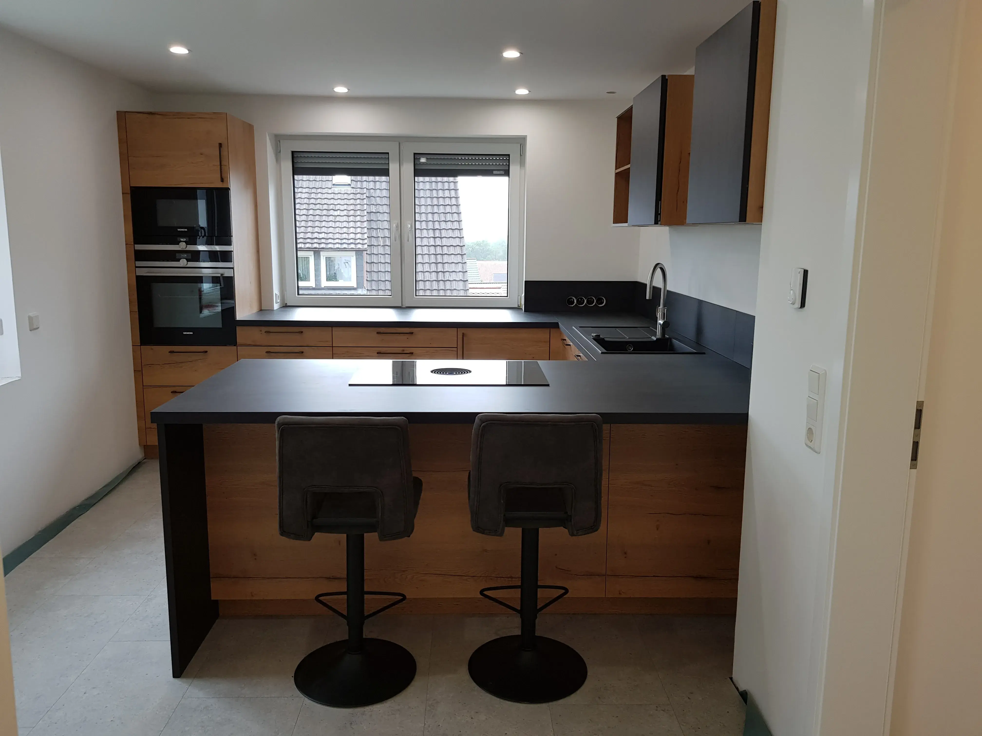 Moderne Küche mit Kochinsel, zwei Barhockern, Holz- und schwarzen Elementen.