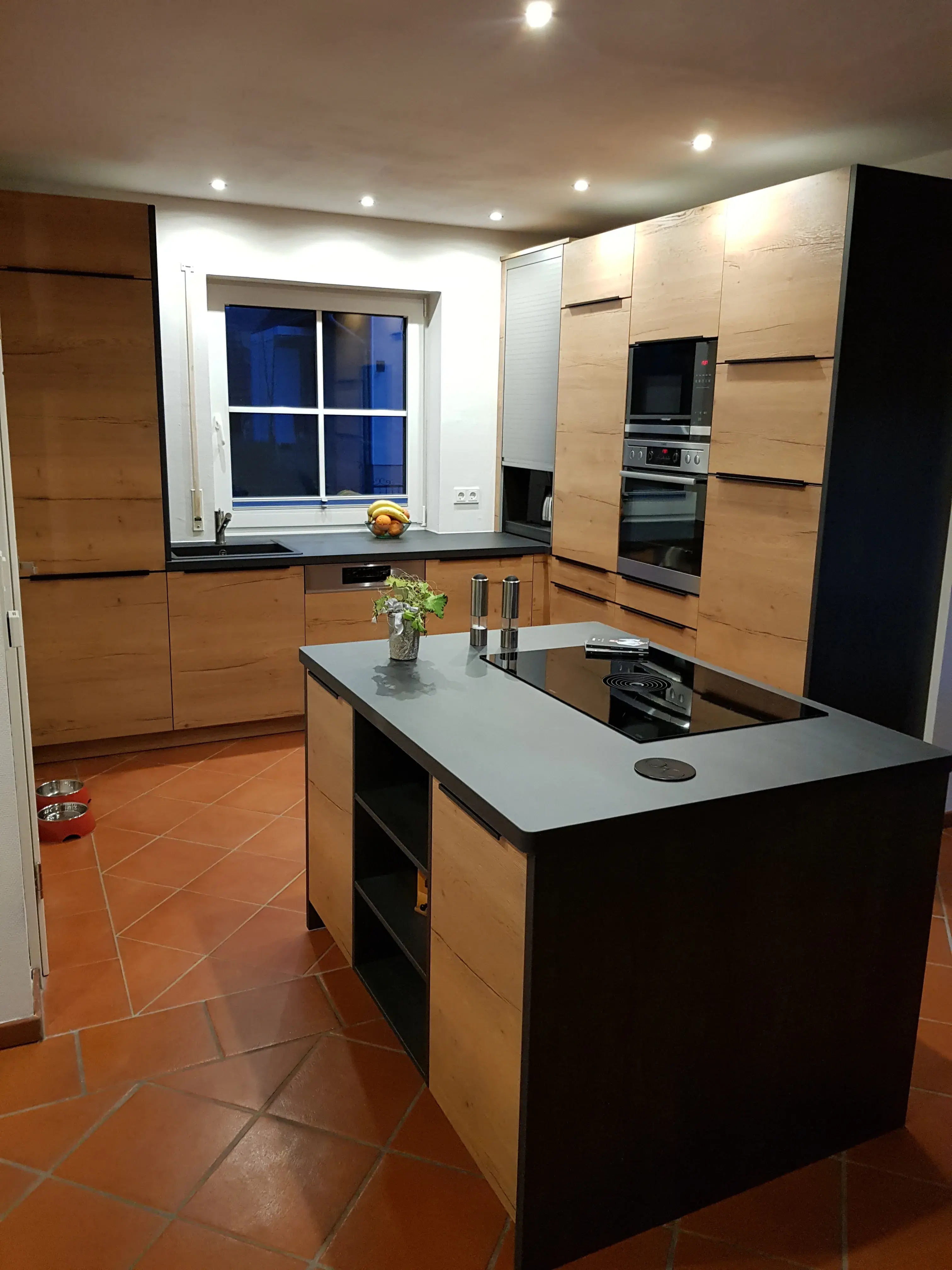 Moderne Küche mit Kochinsel, Holzfronten und grauer Arbeitsplatte, beleuchtet.