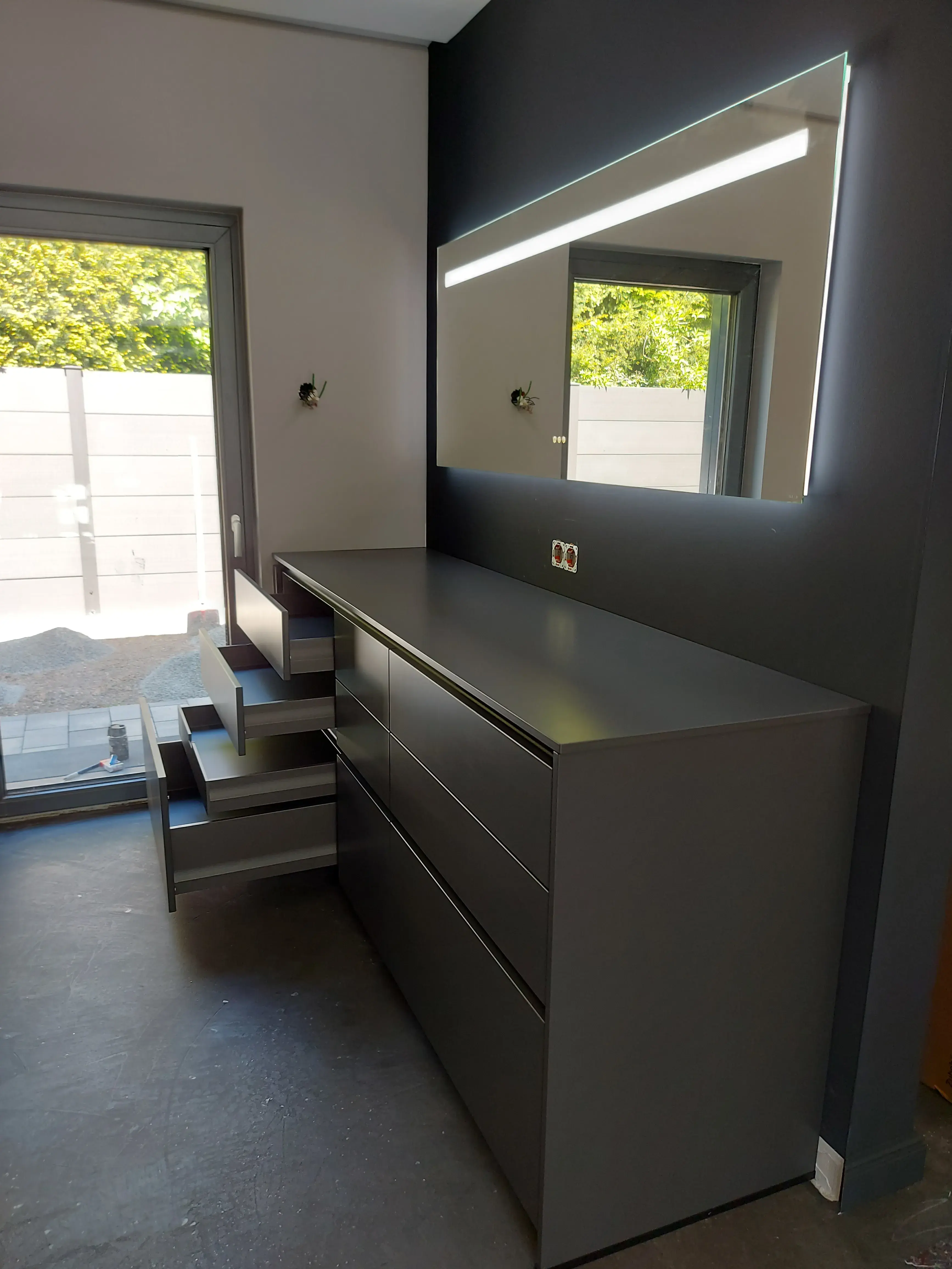 Graue Kommode mit offenen Schubladen vor beleuchtetem Spiegel im modernen Raum.