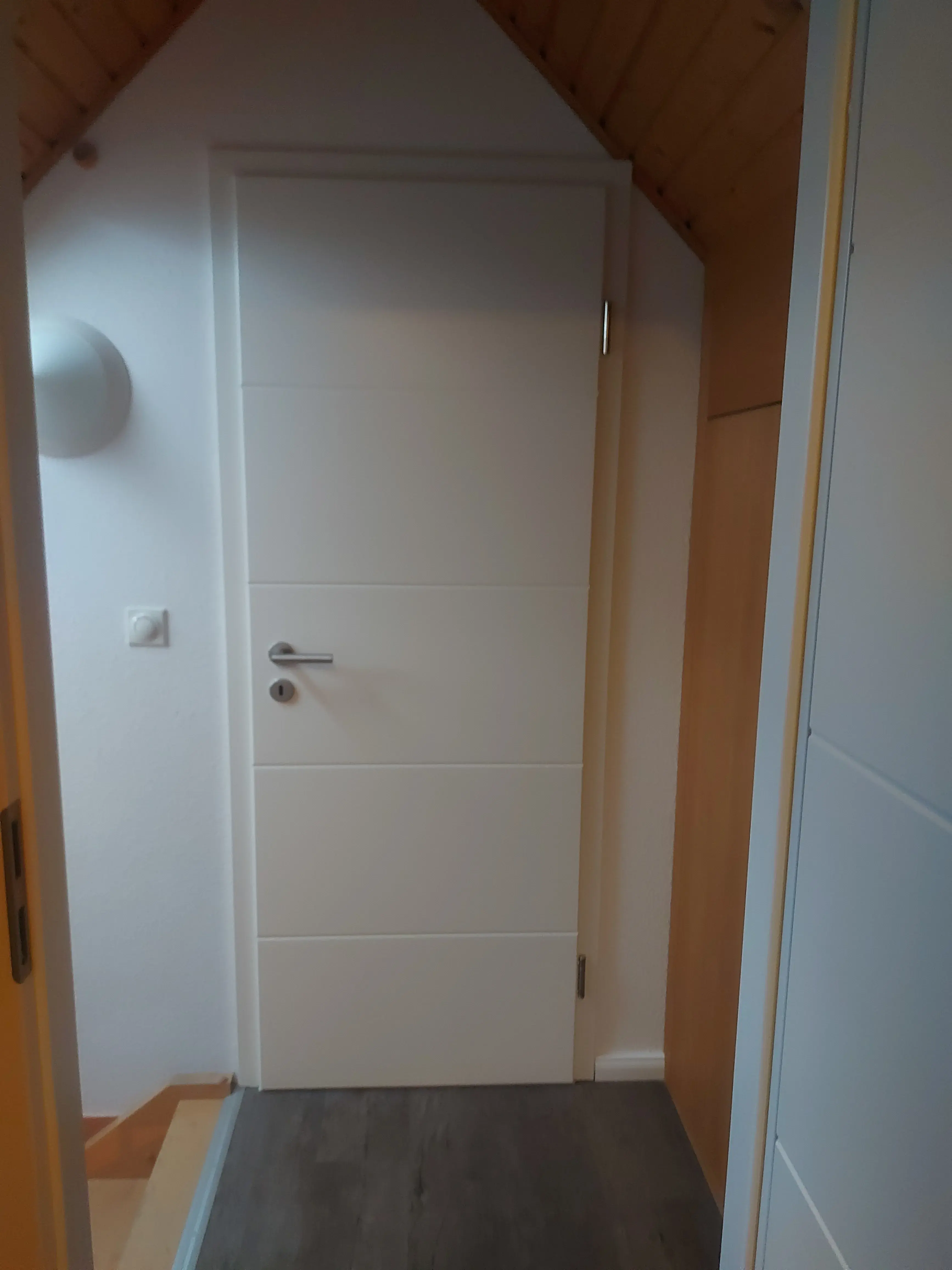 Weiße Zimmertür unter schrägem Holzdach, moderner Flur, grauer Boden.